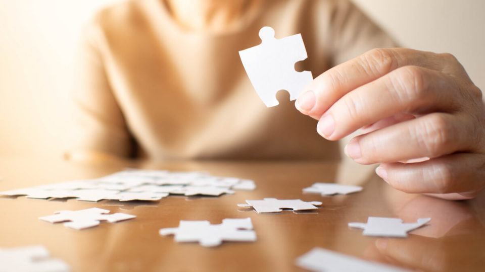 Elderly female hands trying to connect pieces of white jigsaw puzzle on wooden table. Creative idea for Alzheimer's disease, dementia, memory loss