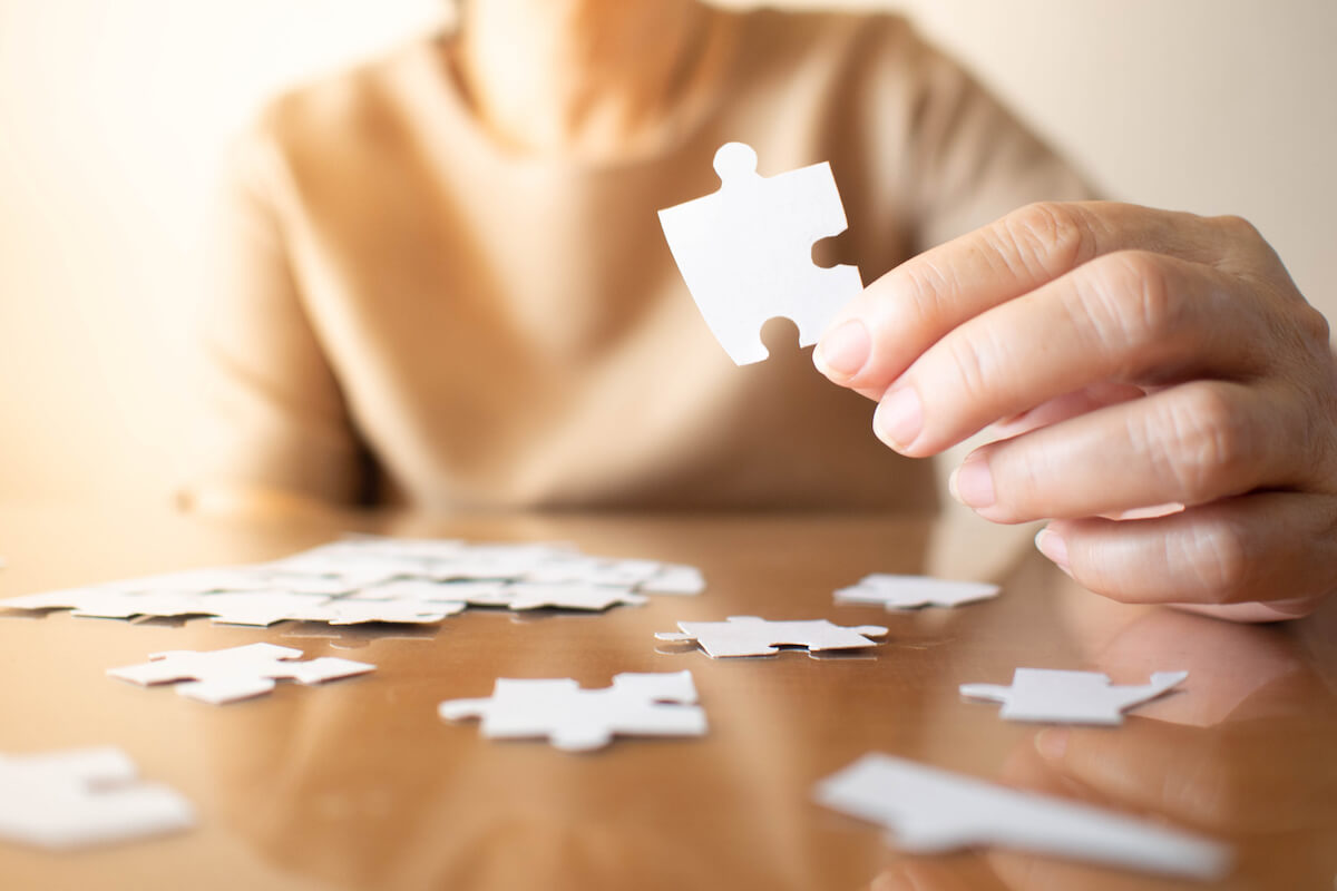 Elderly female hands trying to connect pieces of white jigsaw puzzle on wooden table. Creative idea for Alzheimer's disease, dementia, memory loss